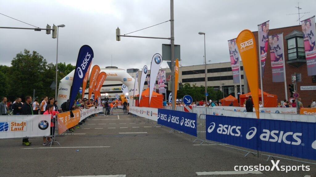 Eine Aufnahme eines Stadtlauf-Events mit Absperrgittern und Werbebannern von Marken wie Asics, BMW und SportScheck. Menschen stehen an den Seiten und warten in der Nähe des Ziels, das durch einen aufblasbaren Torbogen gekennzeichnet ist. Verschiedene bunte Fahnen und Plakate schmücken die Strecke, während das Wetter bewölkt ist.