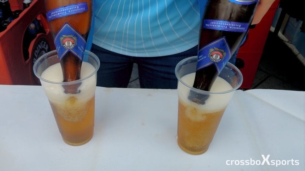 Zwei Gläser mit Bier werden mit Flaschen von alkoholfreiem Erdinger-Weißbier gefüllt. Die Flaschen sind kopfüber in die Gläser gesteckt, während im Hintergrund eine Person mit einem blauen gestreiften Shirt zu sehen ist. Das Bier schäumt leicht am oberen Rand der Gläser